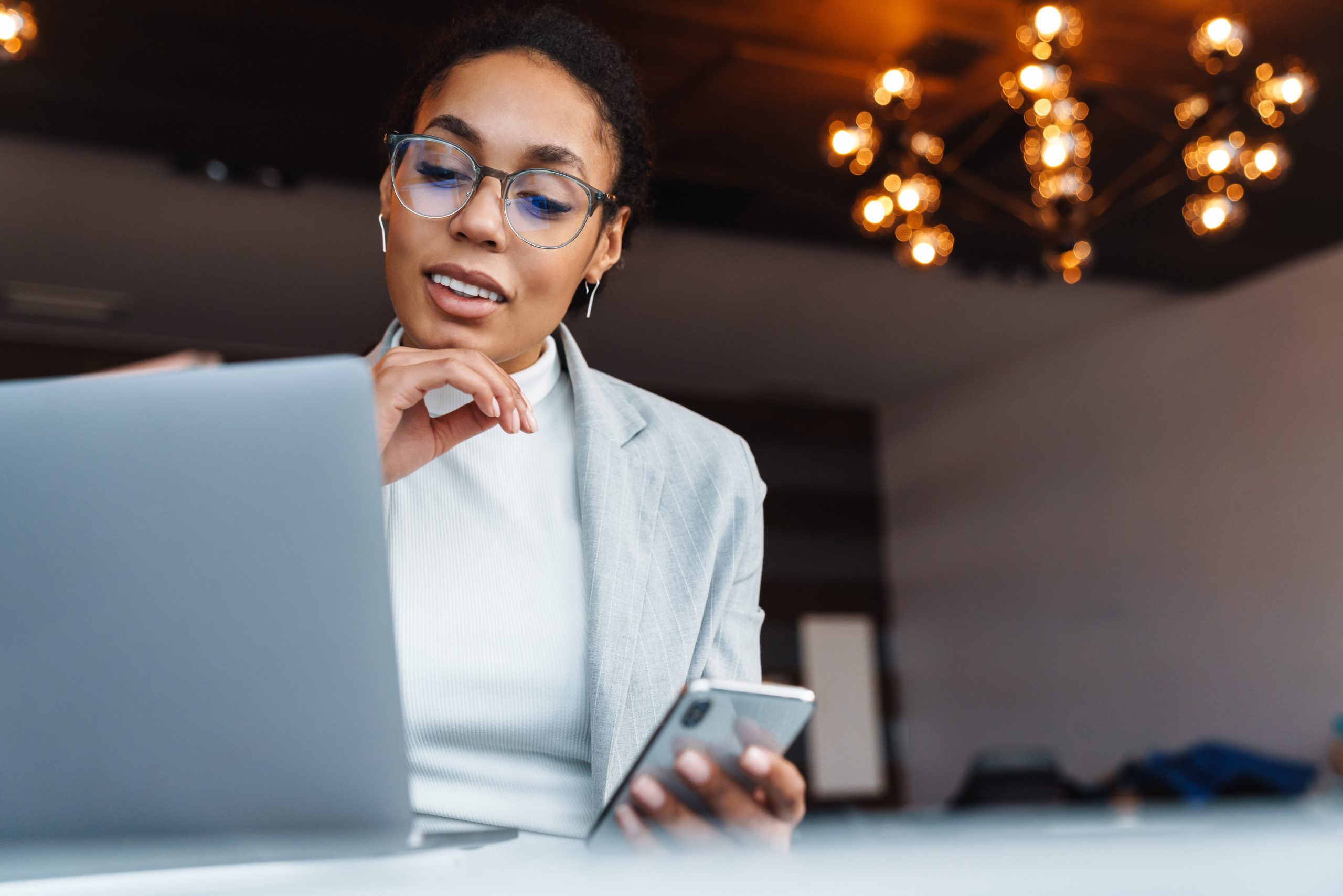 Image of young brunette african american businesswoman using smartphone and working on laptop in office room