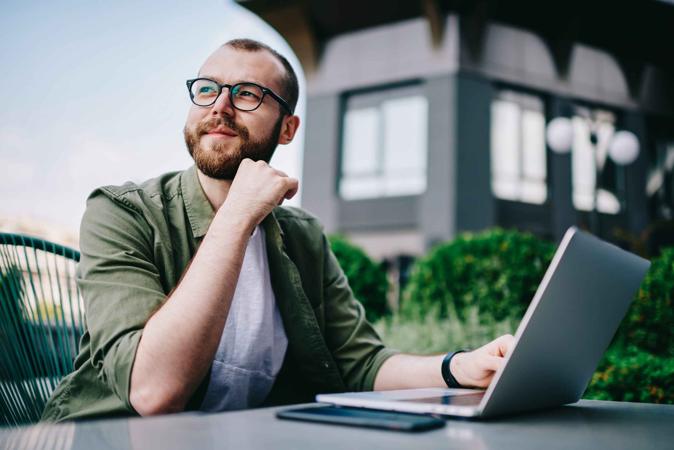 Young handsome male 20 years old in eyeglasses sitting at street cafe during free time with new modern laptop, bearded man freelancer thoughtfully looking aside and thinking about distance work